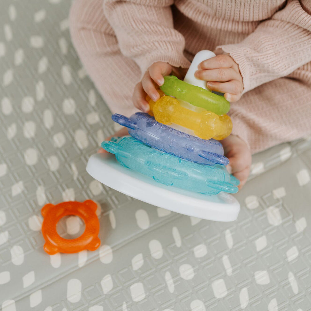 A baby in a pink knit outfit sits on a patterned mat, stacking Nuby Icy Bite Stacking Rings on a white peg base, with an orange teething toy nearby.