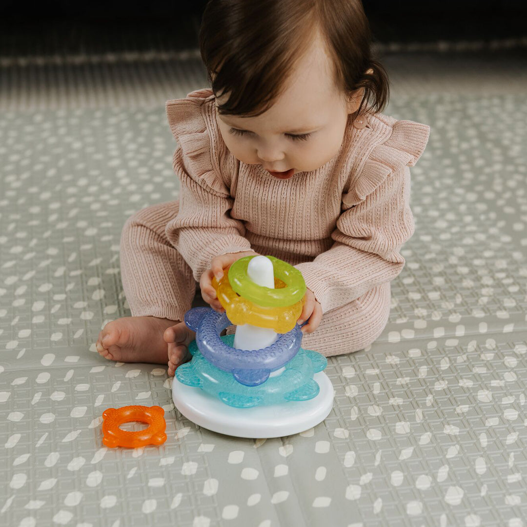 A baby in a pink outfit sits on a patterned mat, playing with Nuby Icy Bite Stacking Rings. She places a yellow teething ring on the white pole while an orange PureICE gel-filled ring rests beside the toy.