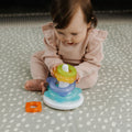 A baby in a pink outfit sits on a patterned mat, playing with Nuby Icy Bite Stacking Rings. She places a yellow teething ring on the white pole while an orange PureICE gel-filled ring rests beside the toy.