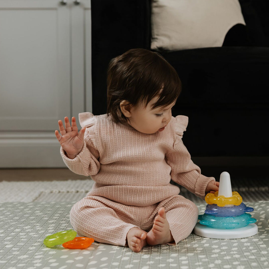 A baby in a pink outfit sits on a patterned rug, playing with Nuby Icy Bite Stacking Rings, a colorful teething toy. The baby reaches for the rings while a black couch and white cabinet are visible in the background.