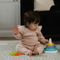 A baby in a pink outfit sits on a patterned rug, playing with Nuby Icy Bite Stacking Rings, a colorful teething toy. The baby reaches for the rings while a black couch and white cabinet are visible in the background.