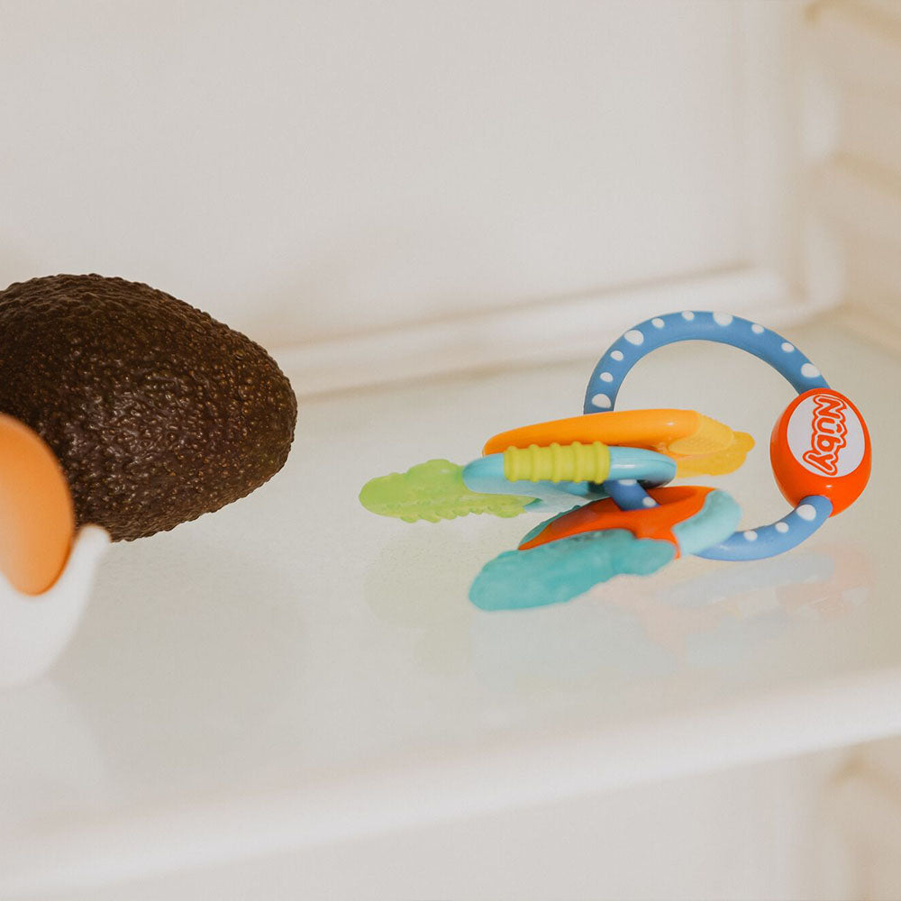 A Nuby Icy Bite Keys Teether Toy, featuring colorful textured keys, is placed beside a half avocado and an egg on a refrigerator shelf.