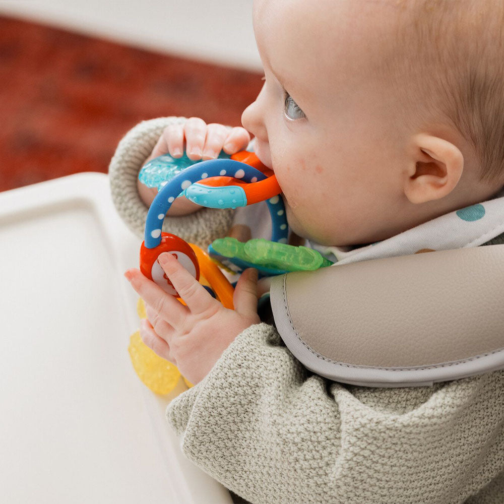 A baby in a high chair holds and chews on the Nuby Icy Bite Keys Teether Toy, a colorful product with various textured loops.
