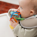 A baby in a high chair holds and chews on the Nuby Icy Bite Keys Teether Toy, a colorful product with various textured loops.