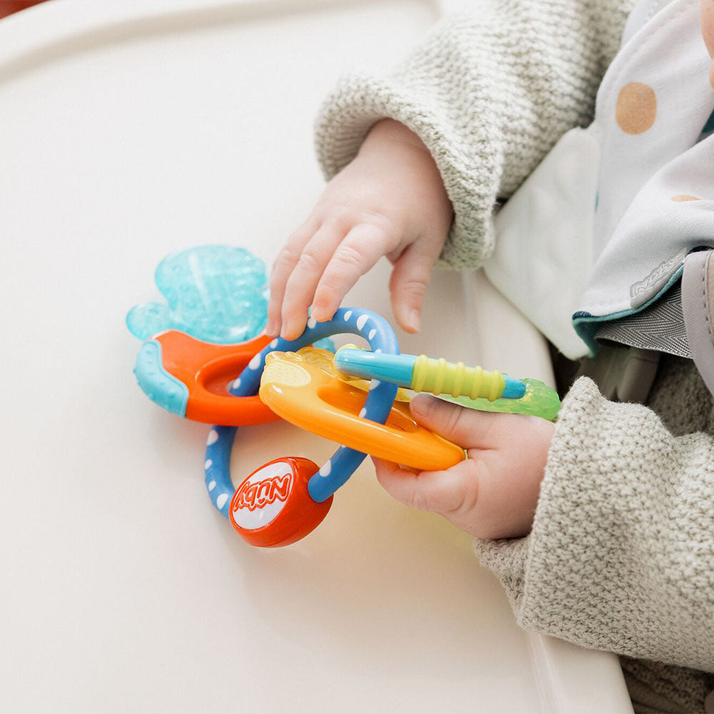 A baby in a gray sweater sits at a white table holding the Nuby Icy Bite Keys Teether Toy, featuring colorful keys with various textures and shapes.