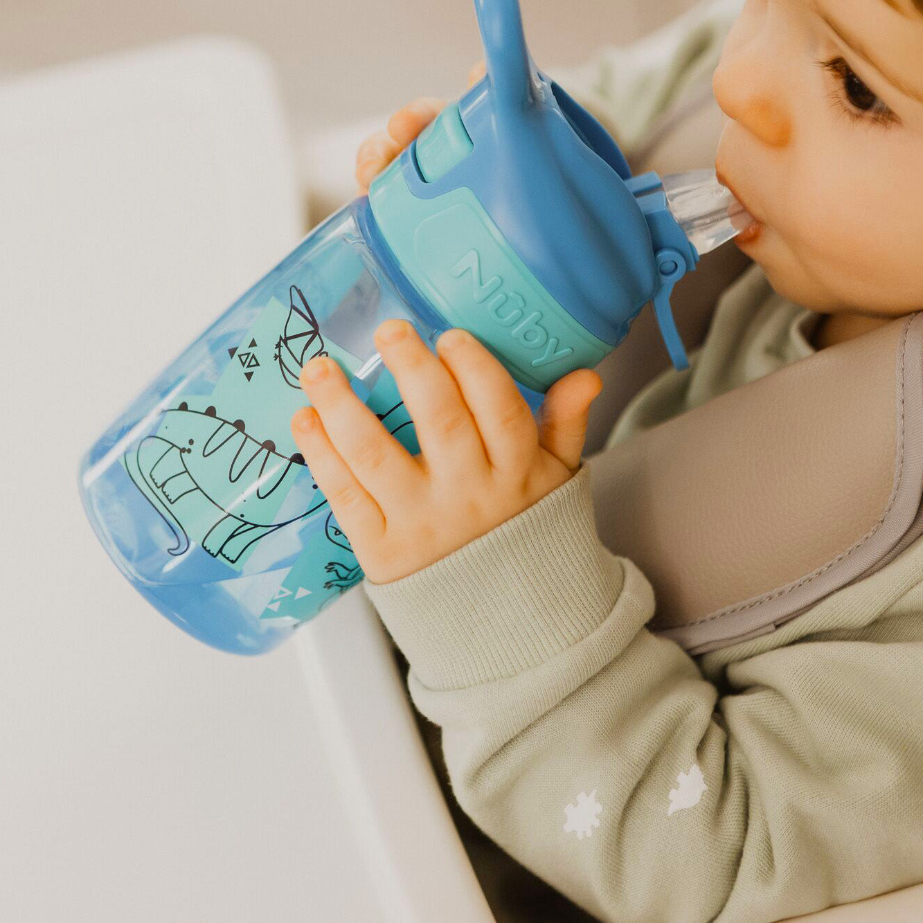 A young child drinking from their Incredible Gulp Dinosaur bottle for babies and young children by Nuby.