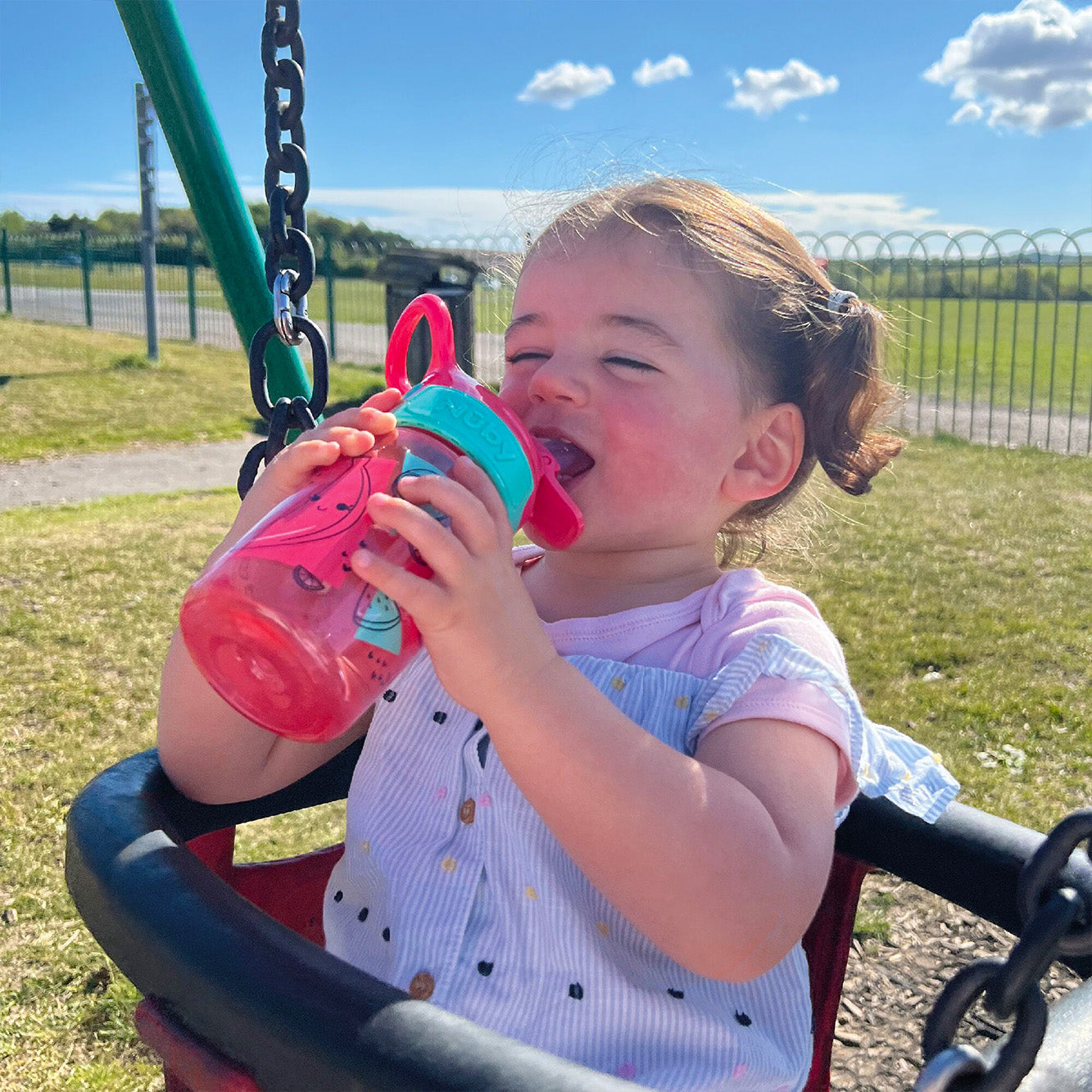 A close up of a young child holding their Incredible Gulp Strawberry decorated bottle by Nuby. They are drinking from the bottle with a smile on their face.