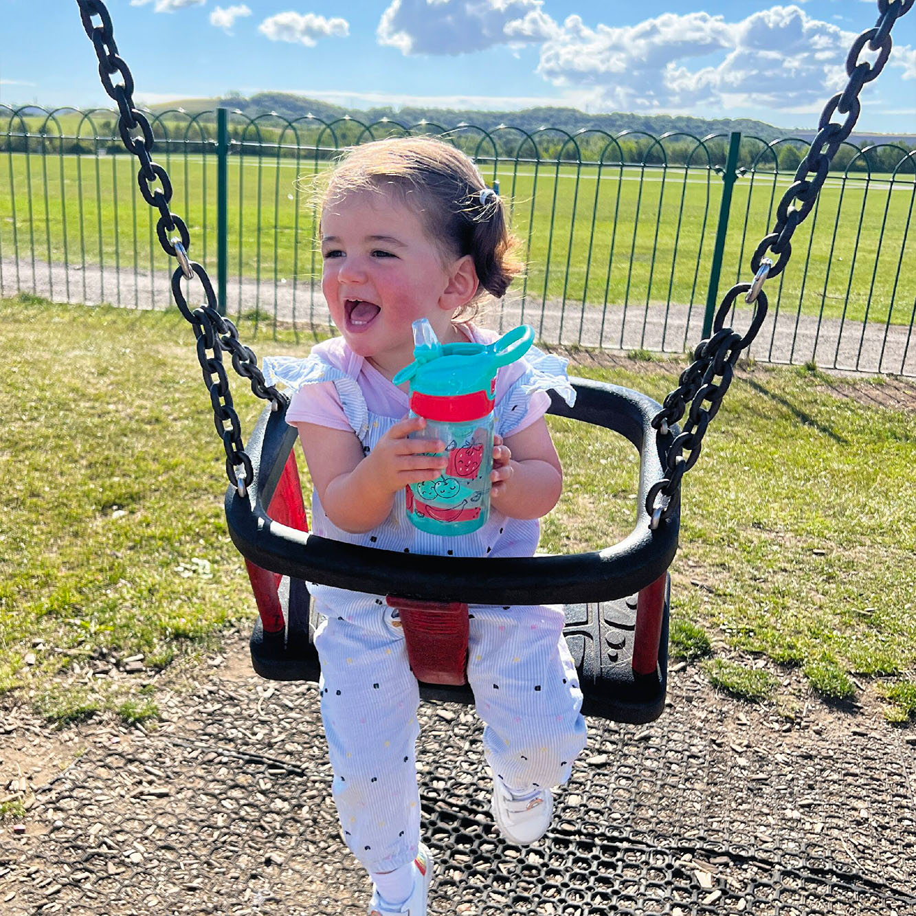 A close up of a young child holding their Incredible Gulp Strawberry decorated bottle by Nuby. They are sat in a swing in a playpark with a smile on their face while carrying the drinks bottle.