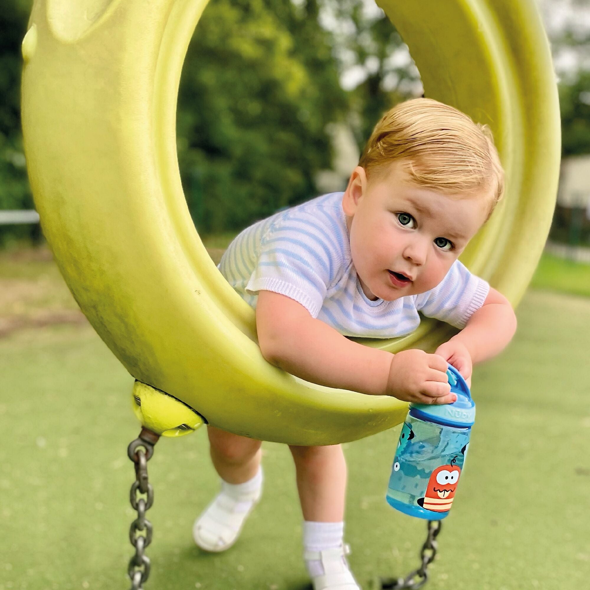 A young boy drinking from the Mighty Swig Monsters sippy cup from Nuby. While playing in the park, in this photo the young boy is holding his cup and looking at the camera.