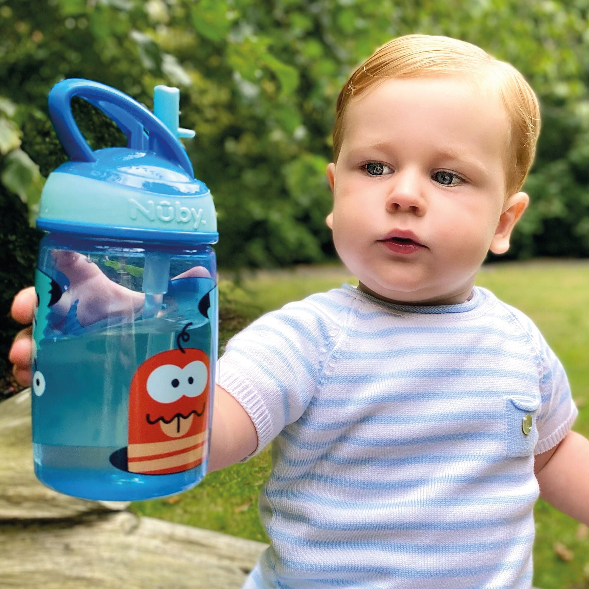 A young boy drinking from the Mighty Swig Monsters sippy cup from Nuby. In this photo he is holding his blue cup and showing it to the camera.