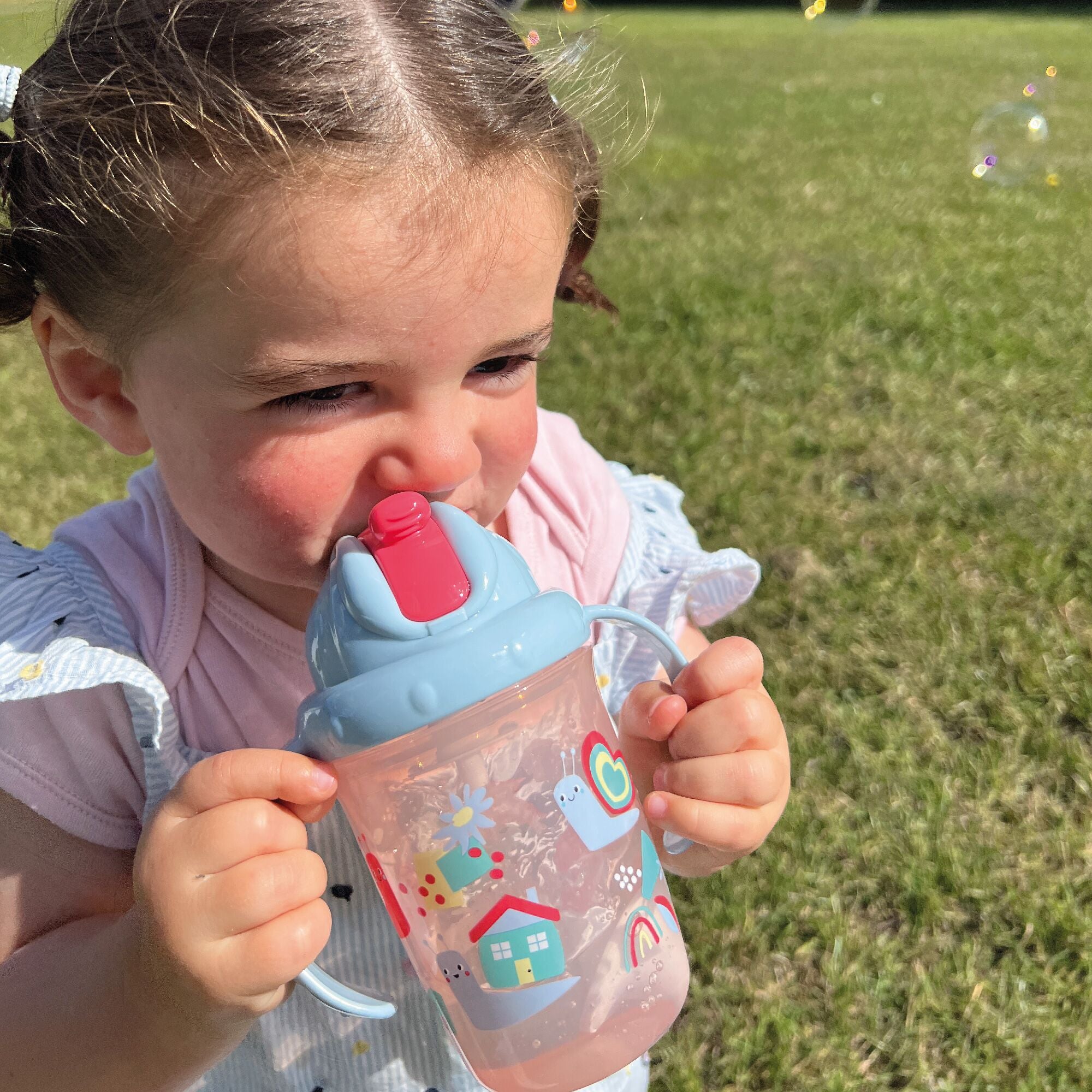 A photo of a young child holding their Flip n Sip sippy cup from Nuby decorated in things from nature. This version has a red straw and snails around the outside of the cup.
