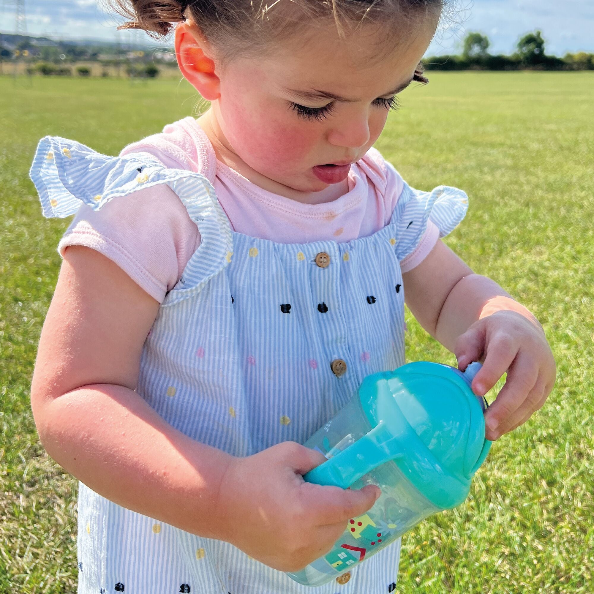 A photo of a young child holding their Flip n Sip sippy cup from Nuby decorated in things from nature. She is using the straw in this image.