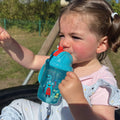 A photo of a young child holding their Flip n Sip sippy cup from Nuby decorated in monsters and in the colour aqua with a red straw. There are friendly monsters waving on the outside of the cup by Nuby.