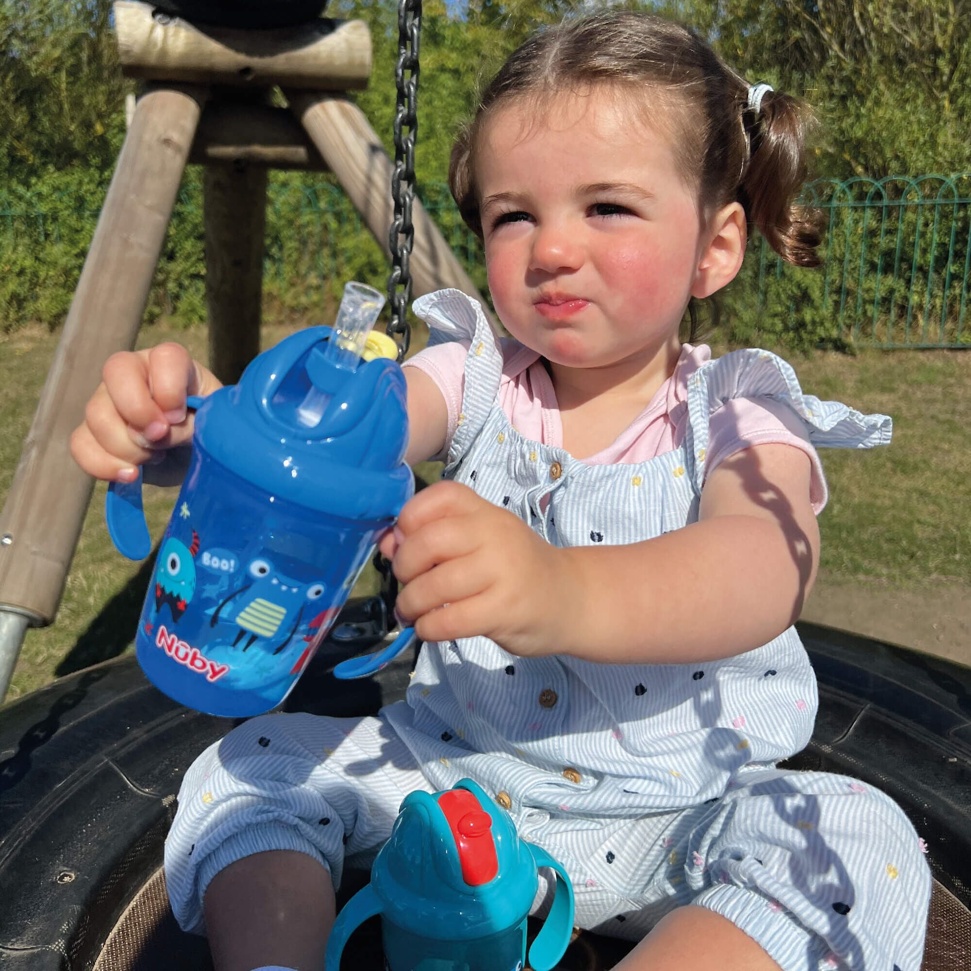A photo of a young child holding their Flip n Sip sippy cup from Nuby decorated in monsters and in the colour blue with a yellow straw. The child is offering to share their drink while sat in a park.