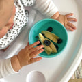 A baby eating banana and blueberries from inside their silicone suction bowl.