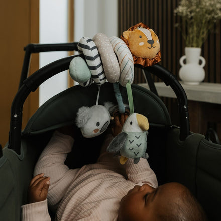 A baby in a stroller reaches for the Nuby Animal Adventures Pram Spiral Activity Toy, featuring lion, koala, and bird characters attached to the handle. A vase with flowers can be seen in the background.