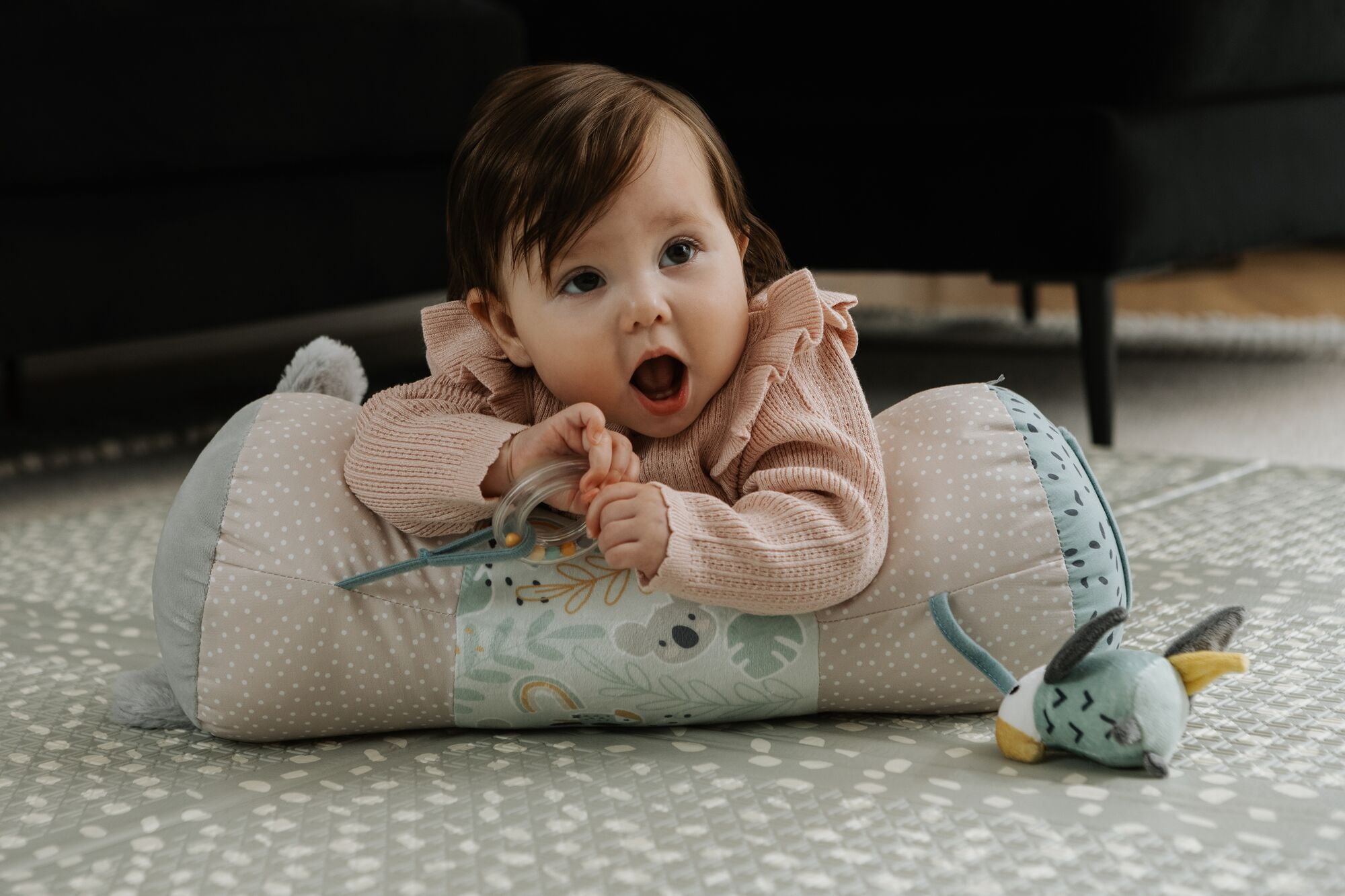 A baby enjoys sensory play on the Nuby Animal Adventures Tummy Time Roller, propped up and clutching a toy, surrounded by playful animal designs and a plush companion.