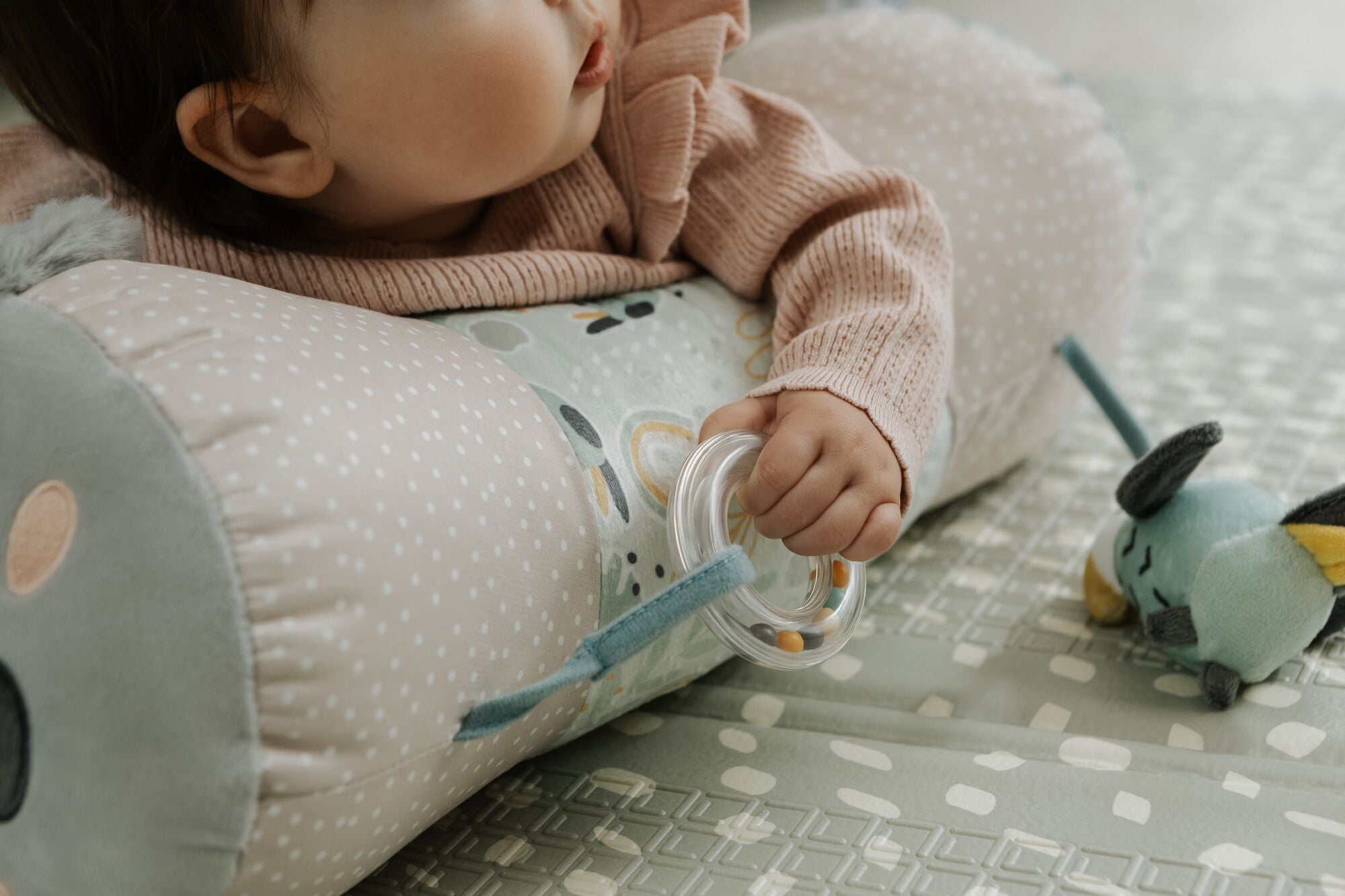 A baby in a pink sweater lies on the Nuby Animal Adventures Tummy Time Roller, holding a clear teething ring. A small plush toy rests on the soft, textured mat nearby, enhancing sensory play.