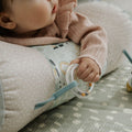 A baby in a pink sweater lies on the Nuby Animal Adventures Tummy Time Roller, holding a clear teething ring. A small plush toy rests on the soft, textured mat nearby, enhancing sensory play.