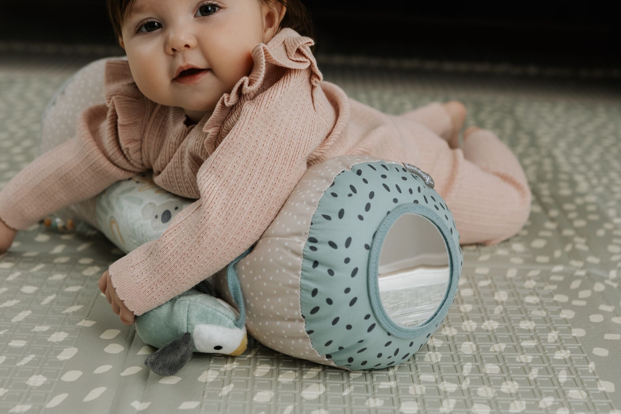 A baby in a pink outfit lies on a patterned mat, hugging the Nuby Animal Adventures Tummy Time Roller with built-in mirror and holding a plush toy—ideal for sensory play and early development.