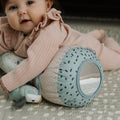 A baby in a pink outfit lies on a patterned mat, hugging the Nuby Animal Adventures Tummy Time Roller with built-in mirror and holding a plush toy—ideal for sensory play and early development.