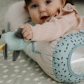 A baby in a pink outfit enjoys sensory play, holding a plush toy and leaning on the Nuby Animal Adventures Tummy Time Roller—a blue, spotted pillow with a circular cutout that offers support during tummy time.