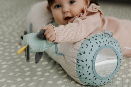 A baby in a pink outfit enjoys sensory play, holding a plush toy and leaning on the Nuby Animal Adventures Tummy Time Roller—a blue, spotted pillow with a circular cutout that offers support during tummy time.