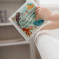 A baby holding their Bathtime Baby Book over the edge of the bath with How many oceans friends can you see on the cover of the book alongside ocean animals.