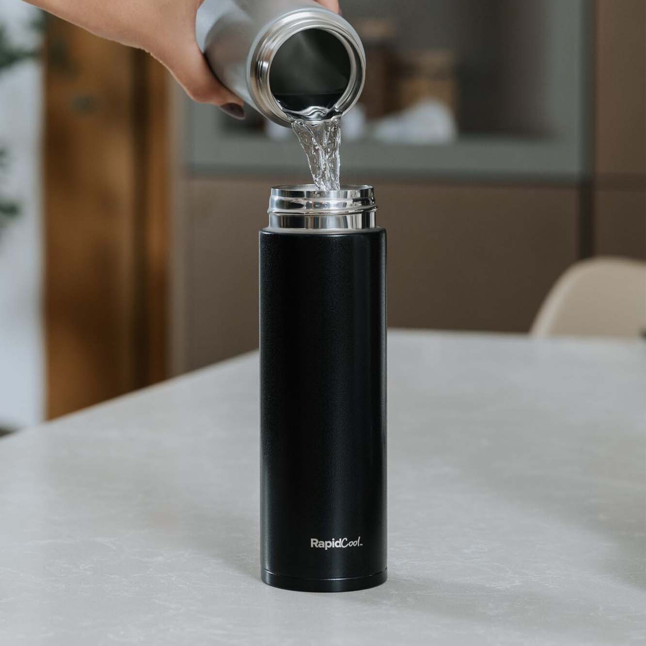 Water being poured into a black RapidCool to help bring the water down to a suitable temperature for a baby to drink from. This is on top of a kitchen table.