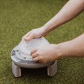A person lines the round, gray Nuby Go Potty with a clear plastic bag on artificial grass, getting it ready for use.