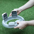 A person’s hands holding a circular gray Nuby Go Potty with three vertical slots, likely used as a toddler travel potty or training seat, on artificial green grass.
