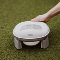 A hand holds the Nuby Go Potty, a portable white and gray potty ideal for potty training, shown on green artificial grass.
