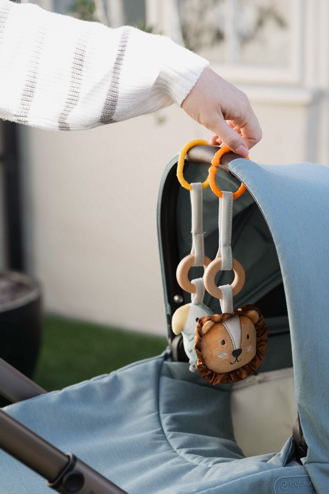 A hand attaches a Nuby Linkables soft toy with plush rings and a lion face to the handle of a blue stroller outdoors, showcasing its durable construction.