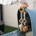 A hand attaches a Nuby Linkables soft toy with plush rings and a lion face to the handle of a blue stroller outdoors, showcasing its durable construction.