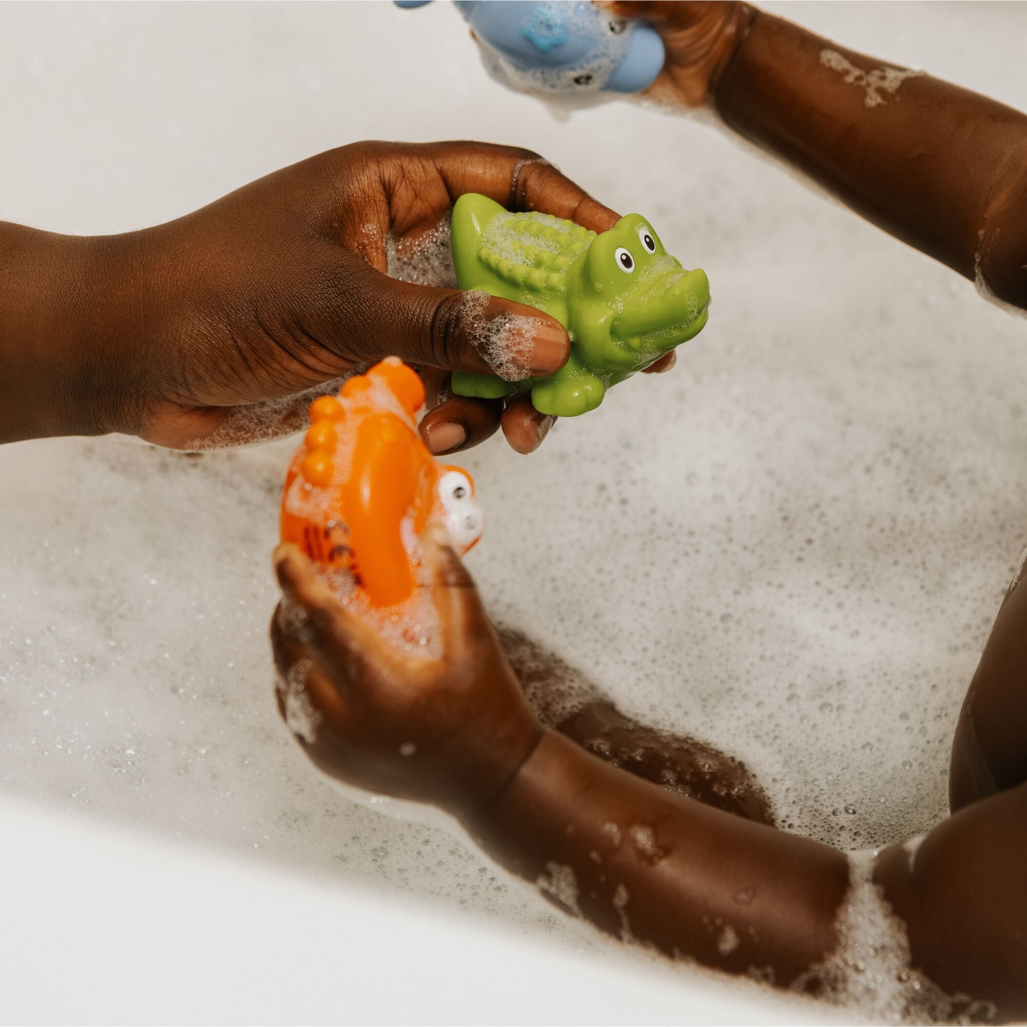 A close-up of two people’s hands holding Nuby Sea Animal Bath Squirting Toys in a bubbly tub—one has a green alligator, the other an orange fish—fun and colorful bath toys perfect for baby’s bath time.