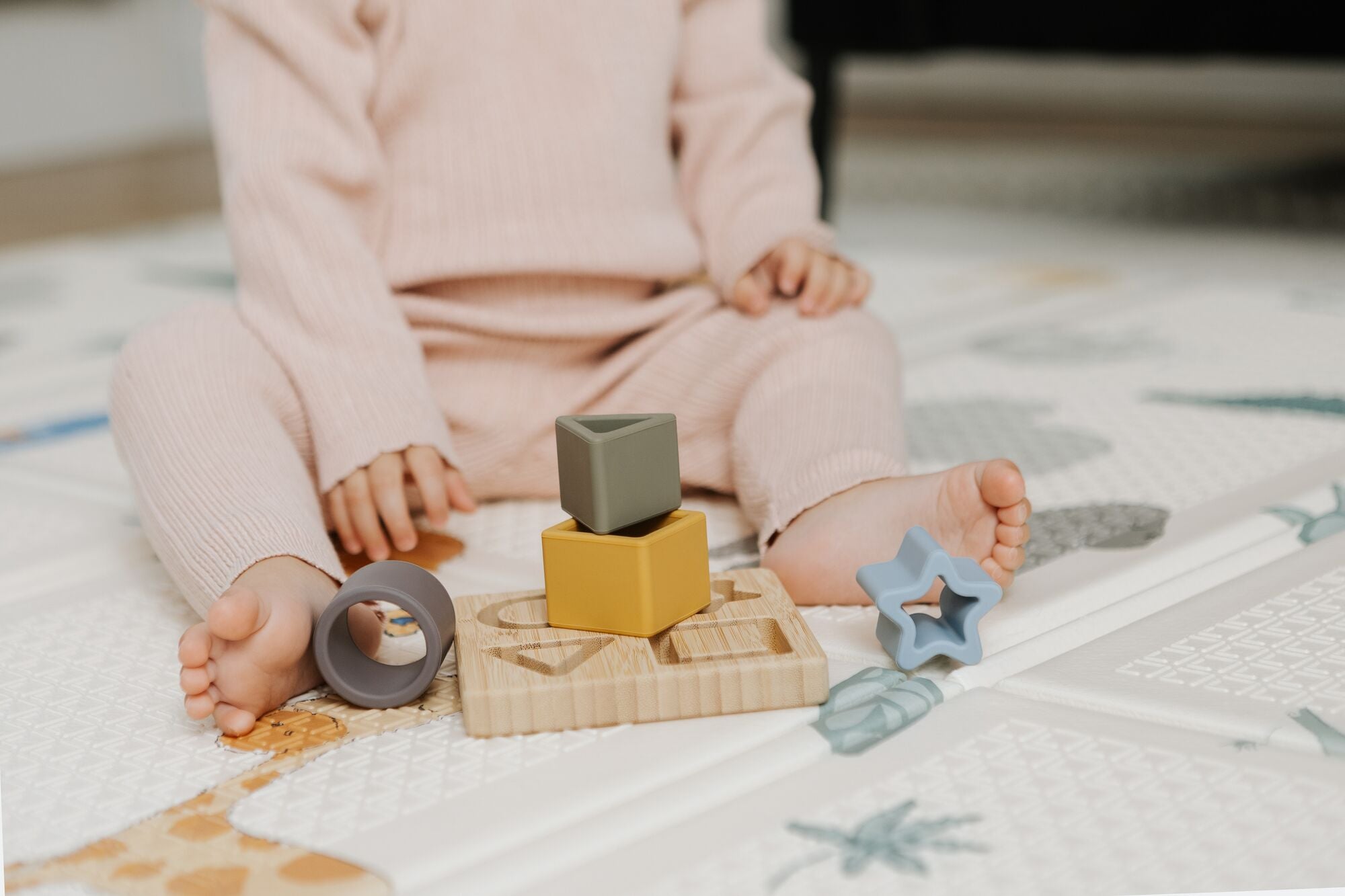 A young child in a light pink outfit sits on a play mat, using the Nuby Shape Sorter Learner Toy with wooden and silicone stacking blocks and rings to build hand-eye coordination, their bare feet visible.