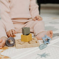 A young child in a light pink outfit sits on a play mat, using the Nuby Shape Sorter Learner Toy with wooden and silicone stacking blocks and rings to build hand-eye coordination, their bare feet visible.