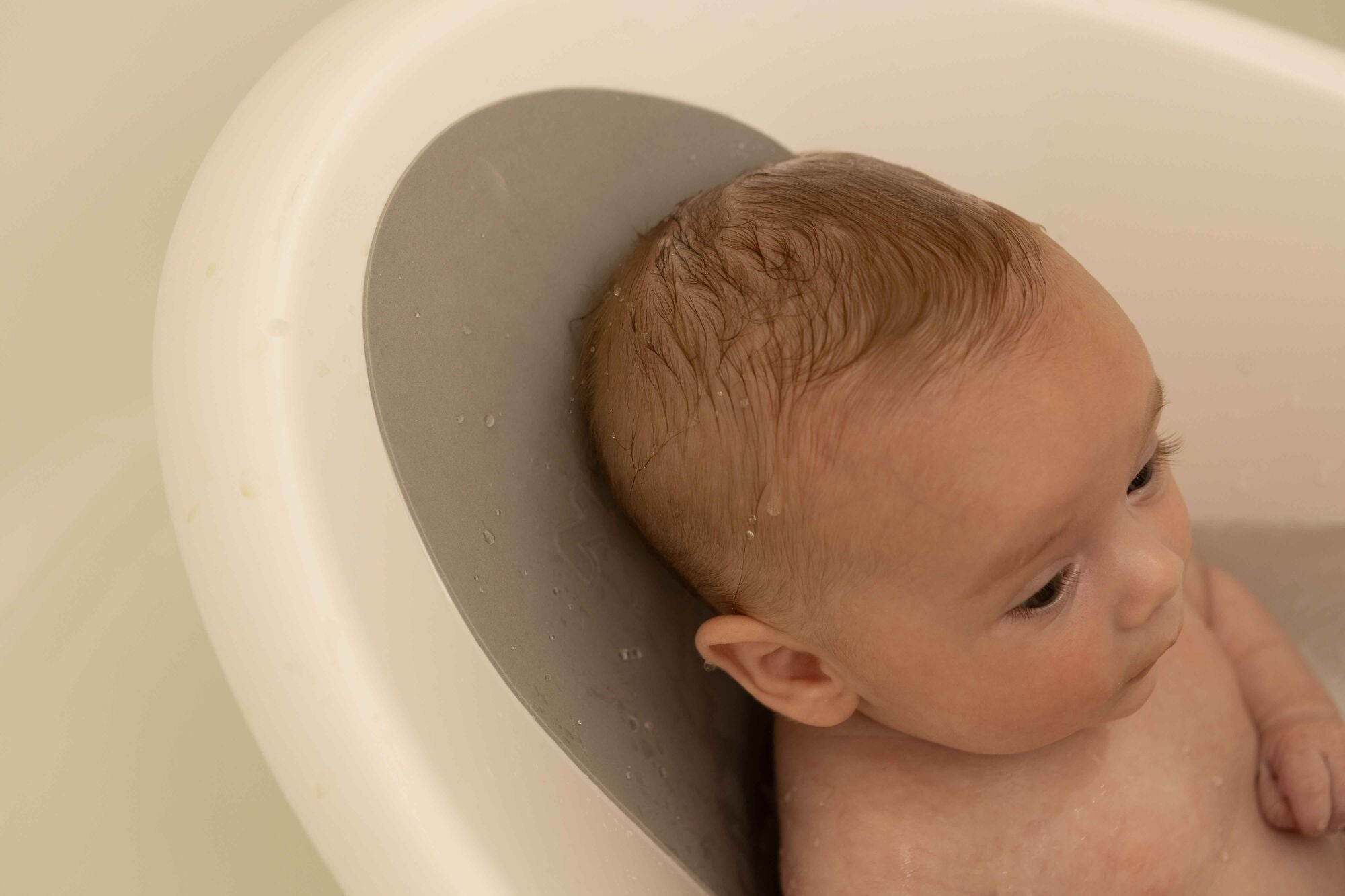 A baby with wet hair relaxes in a white tub, resting its head on the gray Nuby Sit Me Up Baby Bath support. Only the baby's head and upper body are visible as they look off to the side.