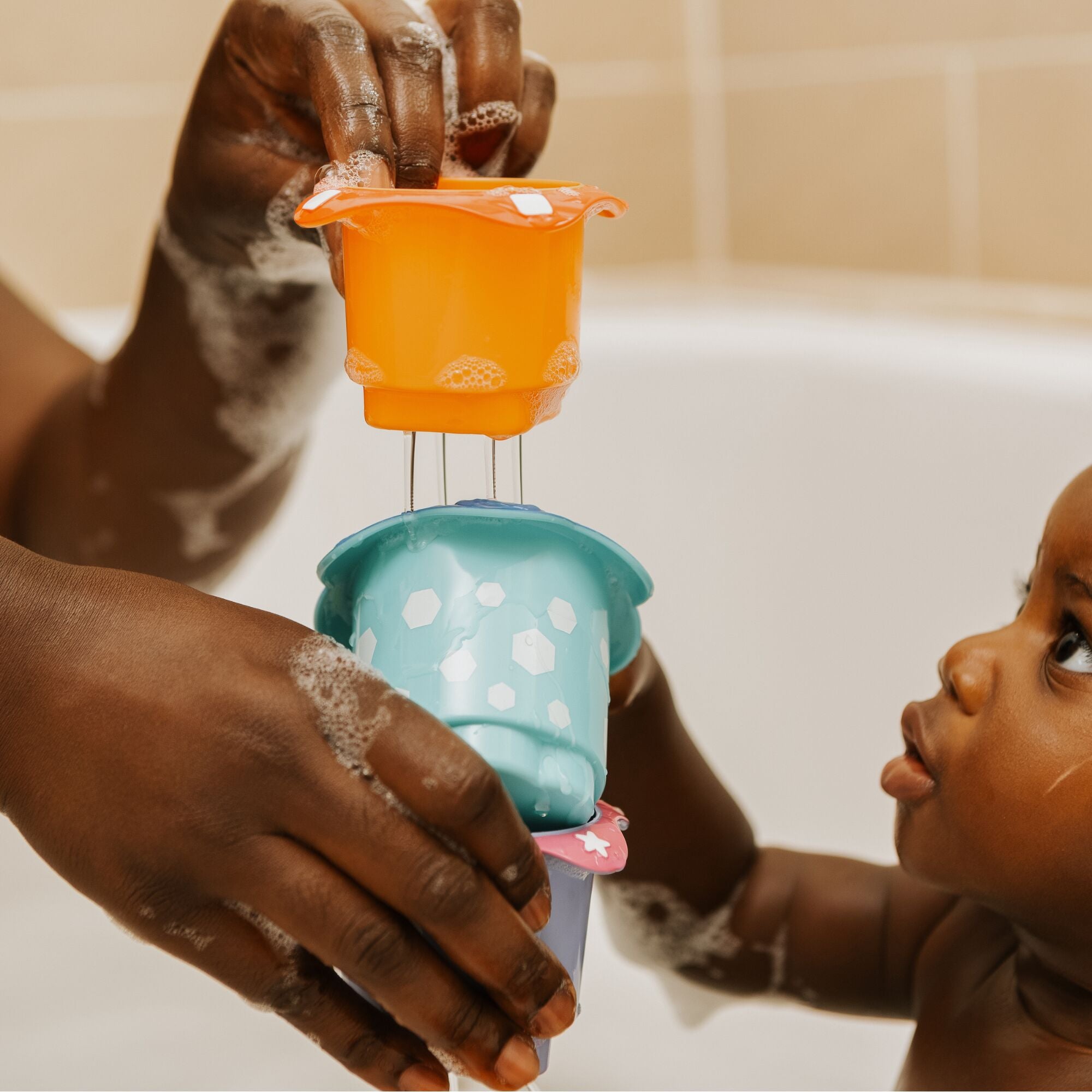 A baby in a bathtub looks up at colorful Splish Splash Stacking Cups by Nuby held by an adult with soapy hands, capturing a playful bath time moment with these fun baby bath toys.