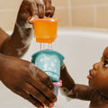 A baby in a bathtub looks up at colorful Splish Splash Stacking Cups by Nuby held by an adult with soapy hands, capturing a playful bath time moment with these fun baby bath toys.