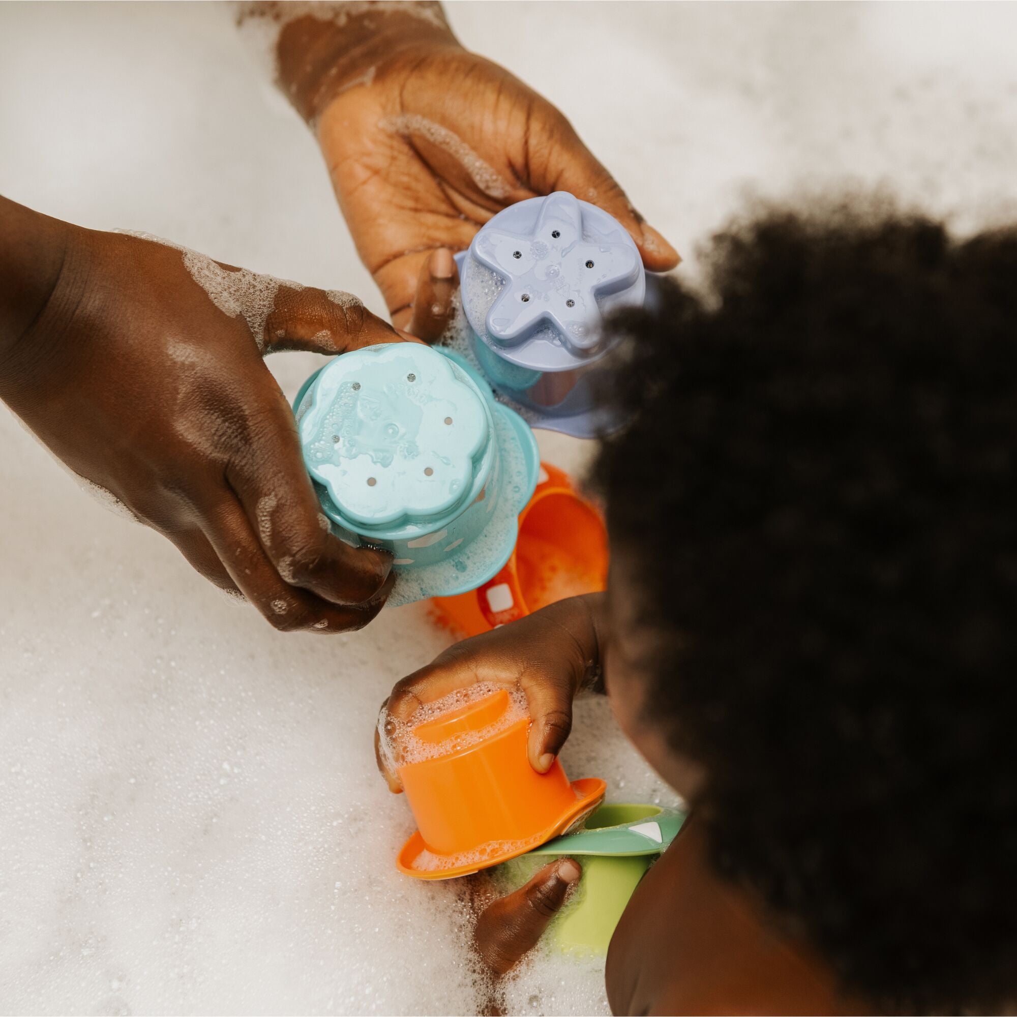 An adult and child enjoy bubbly water play with Nuby Splish Splash Stacking Cups. The adult holds two colorful cups while the child plays with an orange one, showcasing the fun of these baby bath toys.