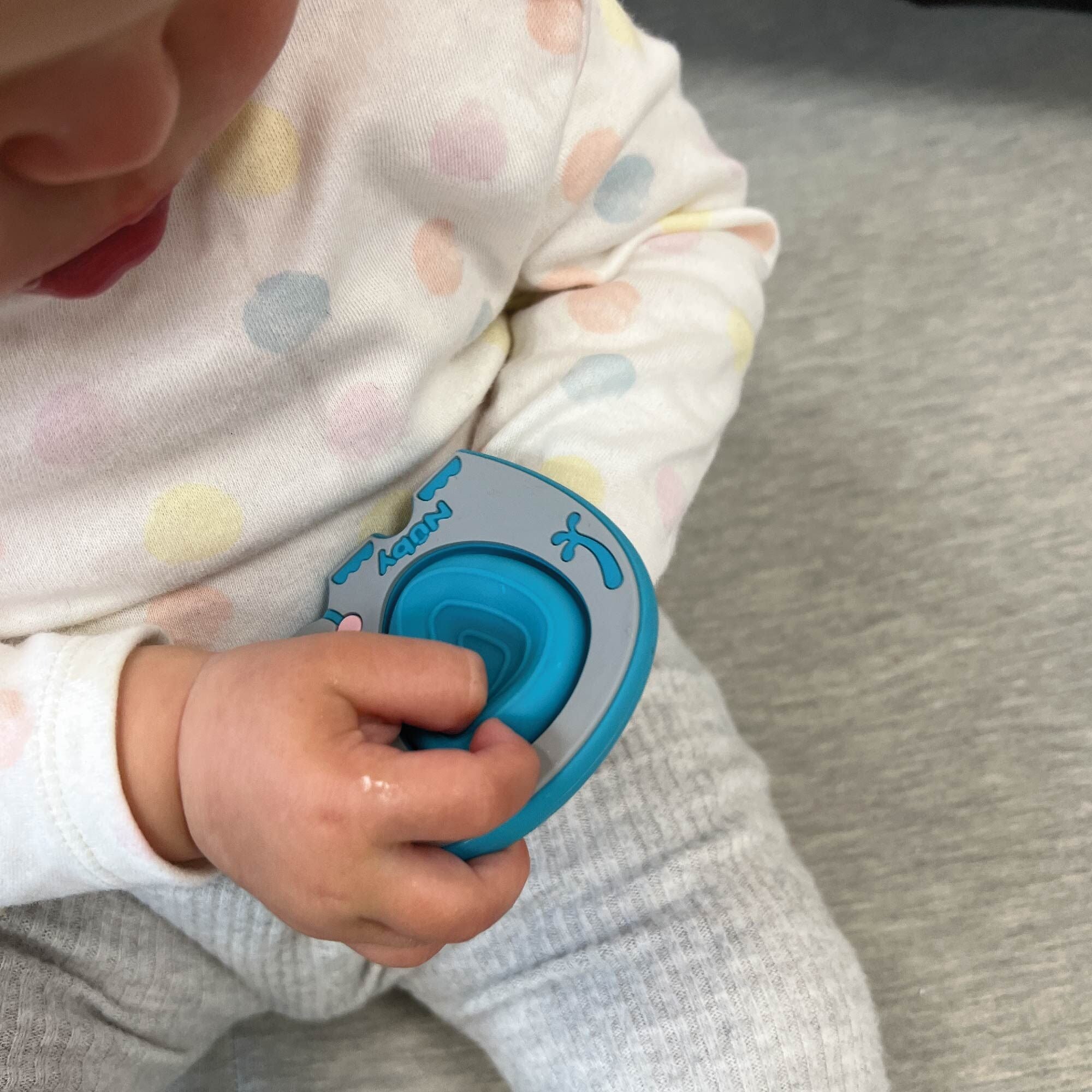 A baby in a white shirt with pastel polka dots and gray pants holds the Nuby Pop-it Teether Toy 2 Pack in blue and gray while sitting on a gray surface, with their face partially visible.