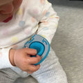 A baby in a white shirt with pastel polka dots and gray pants holds the Nuby Pop-it Teether Toy 2 Pack in blue and gray while sitting on a gray surface, with their face partially visible.