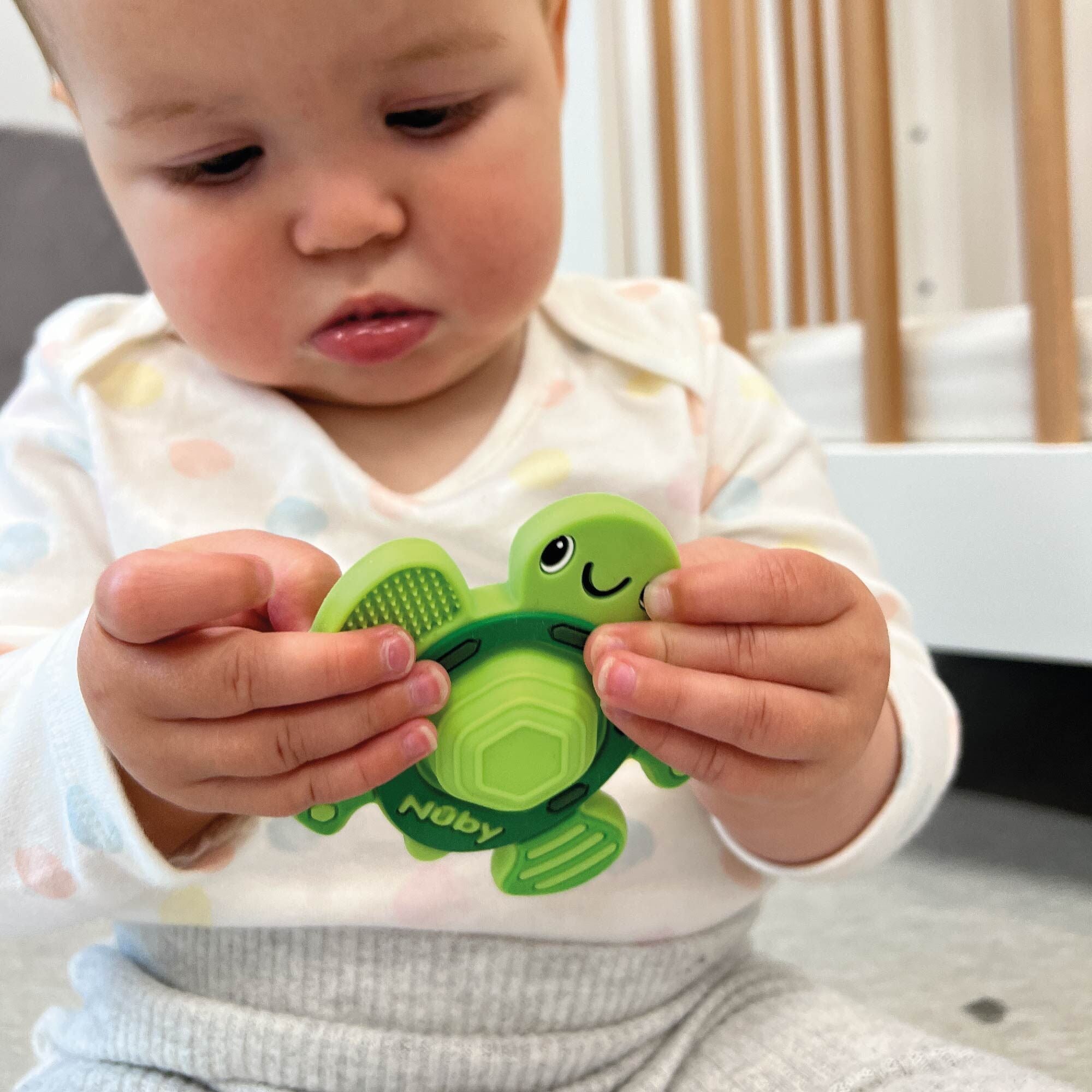 A baby in a white shirt with pastel polka dots holds and examines a green Nuby Pop-it Teether Toy, shaped like a smiling turtle. The background features part of a crib.