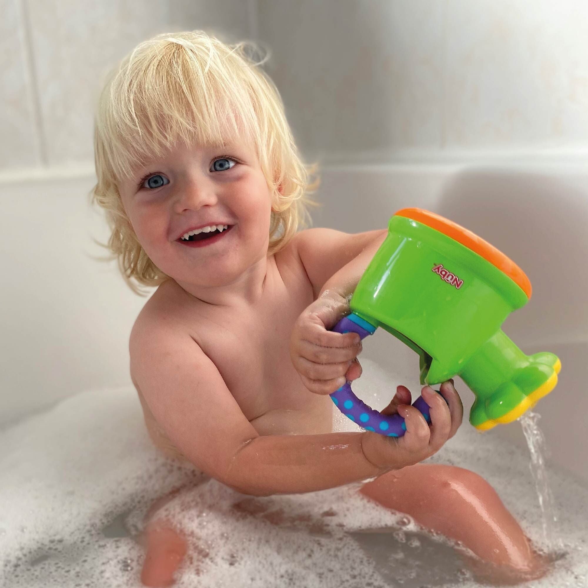 A smiling blonde toddler enjoys bath time with the Nuby Fun Watering Can, a bright green and orange water toy that makes playtime happy and playful.