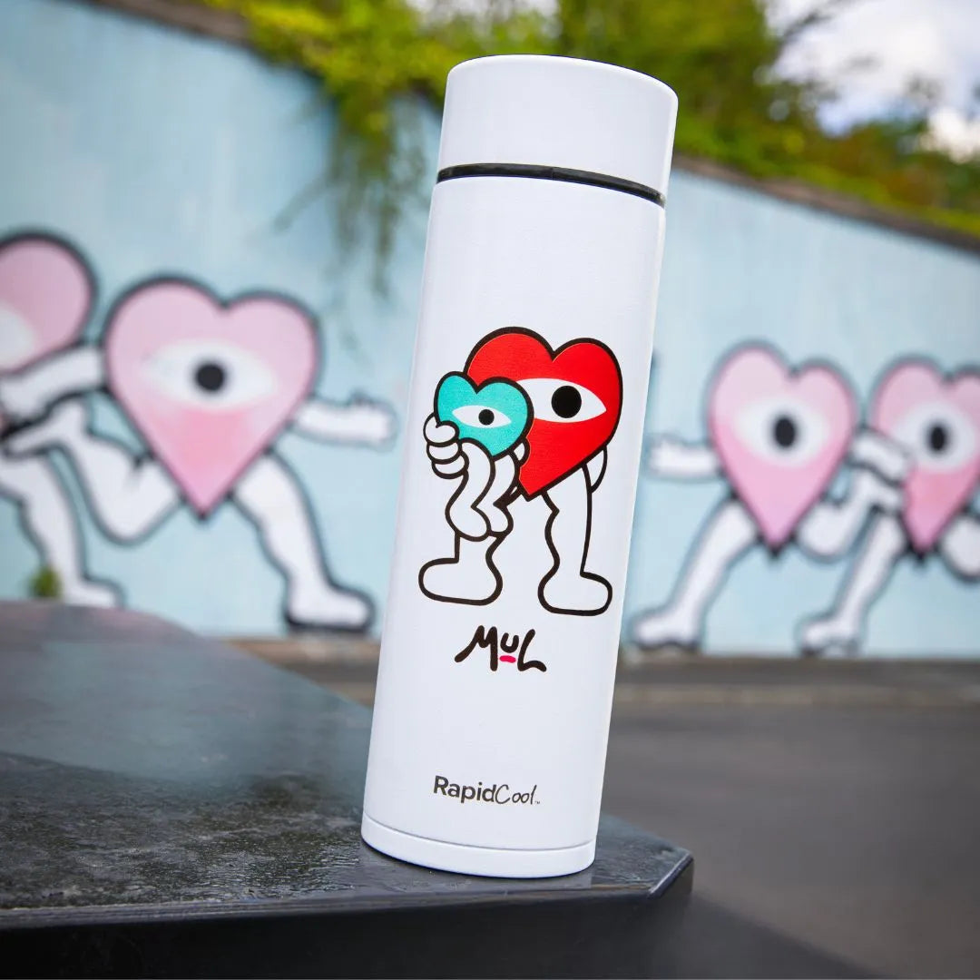 A Limited Edition Nuby RapidCool x Mul white thermos features a cartoon heart holding a blue figure, standing on a black surface, with a mural of similar heart characters on the wall behind it.