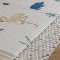 Close-up of the Nuby Foldable Play Mat, Safari, showing its textured white surface with a flamingo, green leaves, and blue shapes on a woven rug over wood.