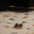 A Nuby Foldable Play Mat, Safari, featuring charming animal illustrations, lies on the floor with a small wooden toy set on top. Soft lighting highlights a partially visible dark-colored sofa in the background.