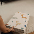 A person places the Nuby Foldable Play Mat, Safari, with colorful animal designs like a giraffe, flamingo, and rhinoceros on a white background, on a textured rug.