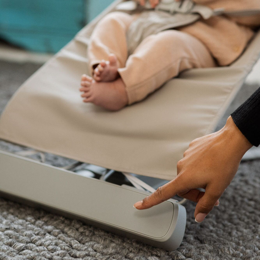 A baby relaxes in the Nuby Musical Moves Baby Bouncer Chair as an adult with a dark sleeve presses a button on the beige frame; the scene is set on a textured grey carpet with the baby's feet visible.